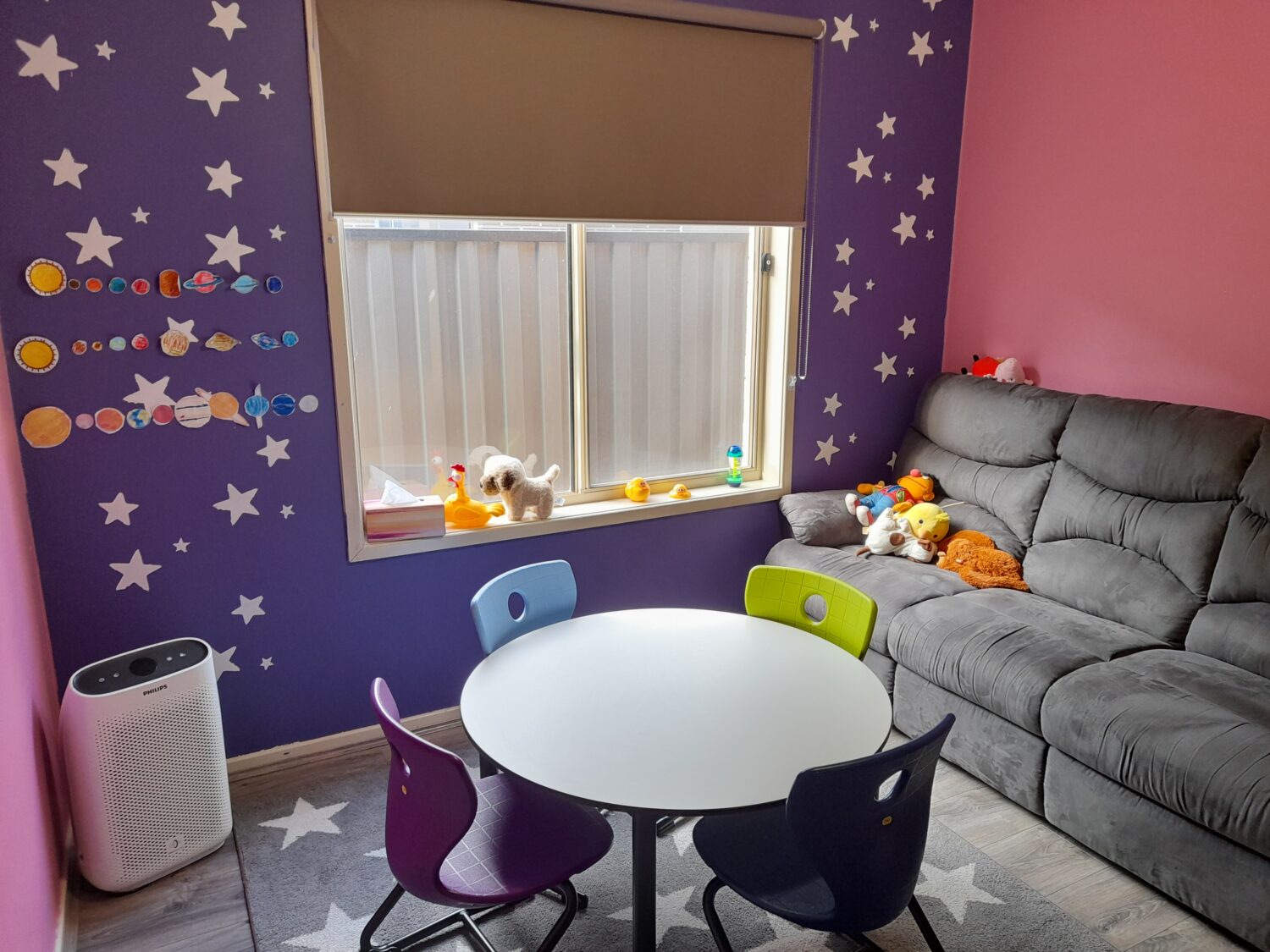 A children's playroom with purple and pink walls, star decorations, a round table with colorful chairs, a couch with stuffed animals, and a window with a few toys on the sill—perfect for transforming into a cozy therapy room rental.