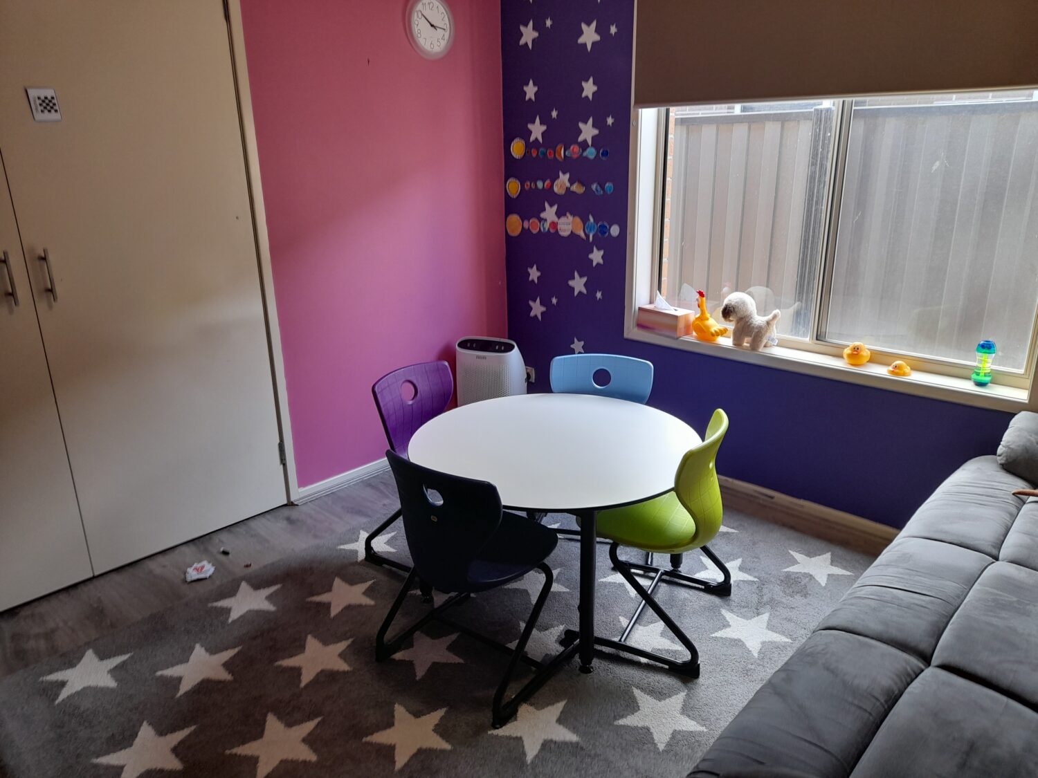 A small playroom with a pink and purple wall, featuring a round white table surrounded by colorful chairs, star-patterned rug, toys on the windowsill, and a closed door with a clock above it—perfect for therapy room rental or those looking to rent therapy office space.