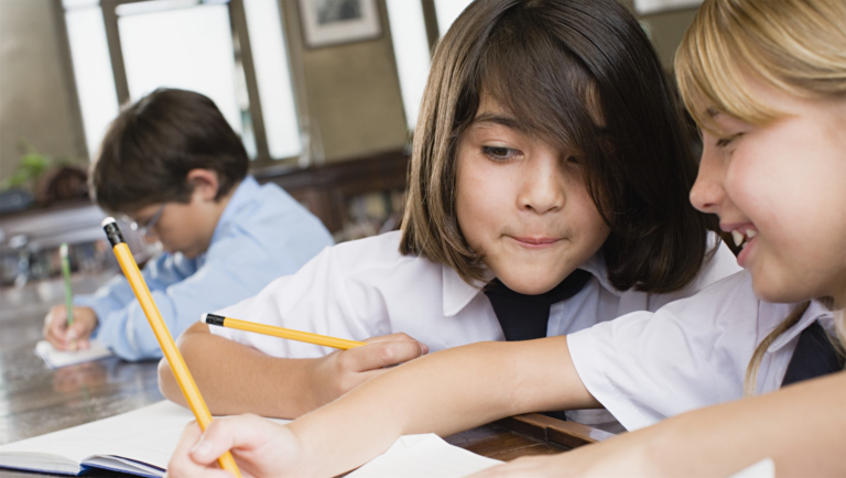 Two children are sitting at a desk and writing in a notebook.