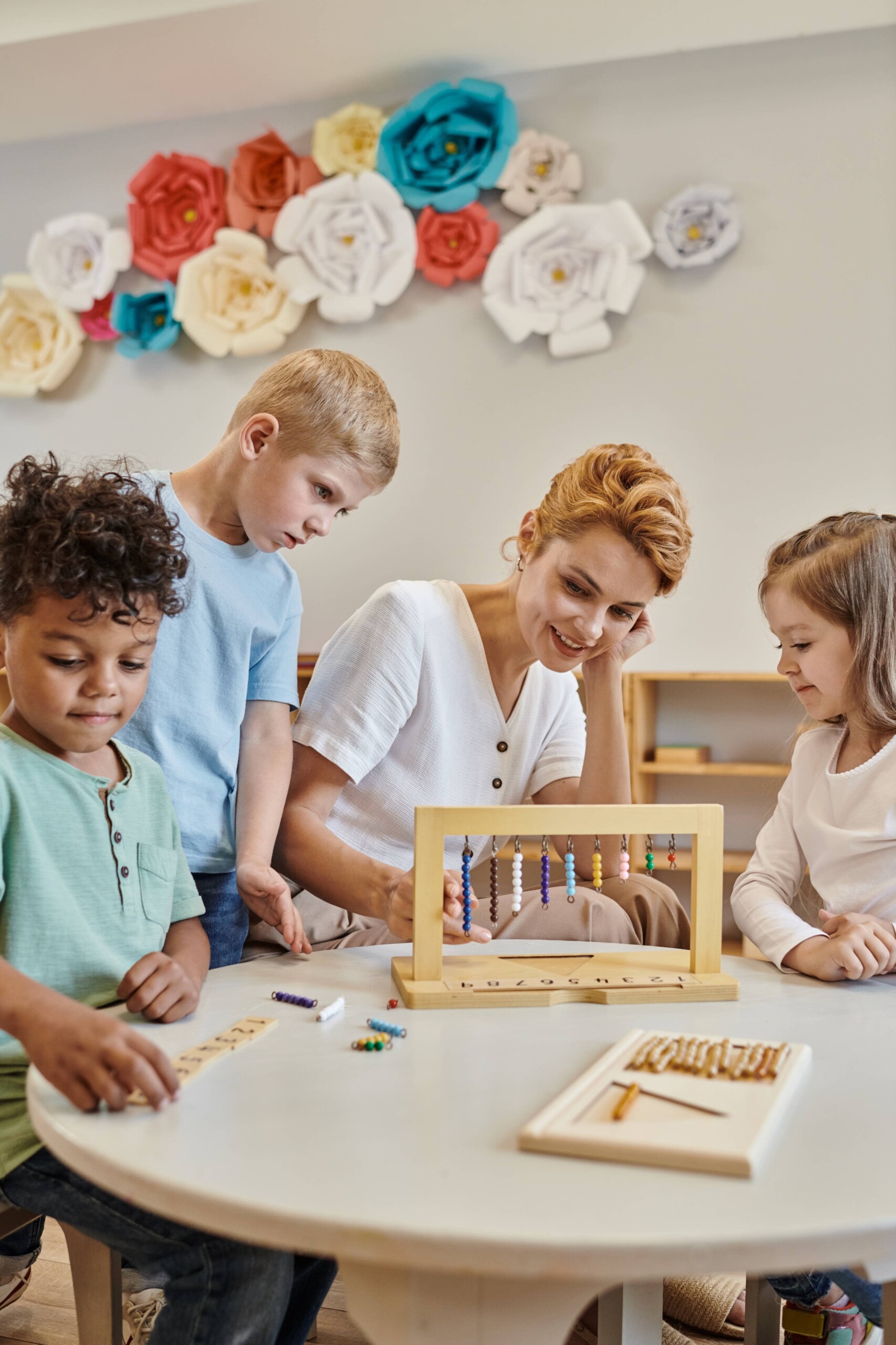 A woman and three children, two boys and a girl, are engaged in a learning activity with an abacus in a decorated classroom.