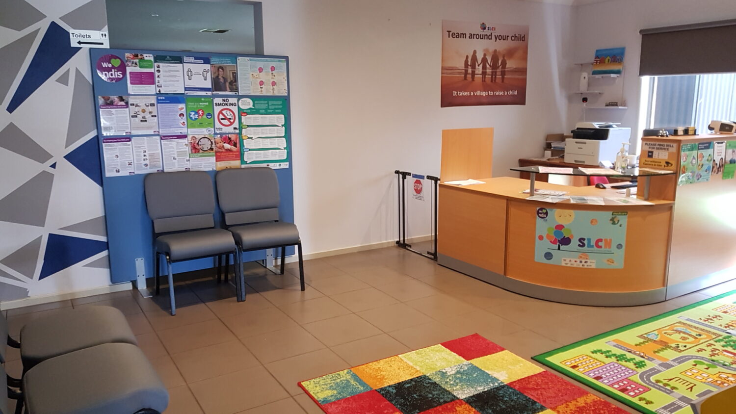 A waiting area with grey chairs, informational posters on the wall, a reception desk, and a children's play area with colorful mats and toys. A sign reads "team around your child. " the space also offers therapy room rental for those in need of a private session space.