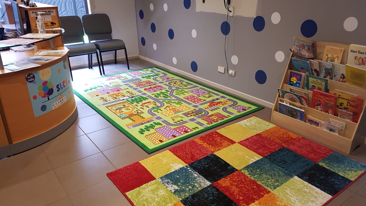 A cozy waiting area features children's play mats, a bookshelf with children's books, two chairs, and a round counter with brochures—an ideal setup if you rent therapy space for your practice.