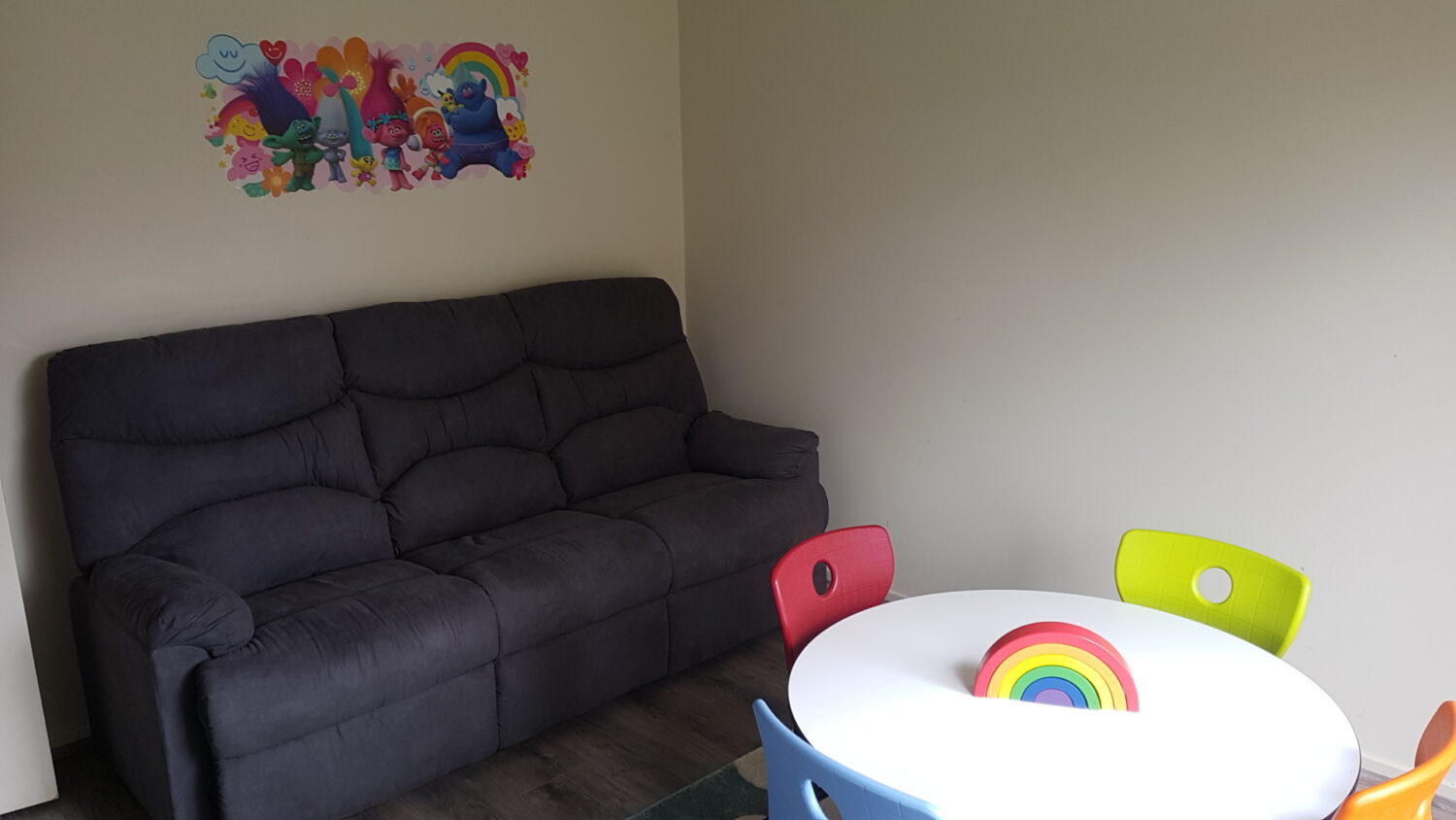 A small therapy room with a dark grey couch, a white round table with four colorful chairs, and a rainbow decoration. A poster featuring cartoon characters is on the wall above the couch. Perfect for those looking to rent therapy space in a cozy setting.