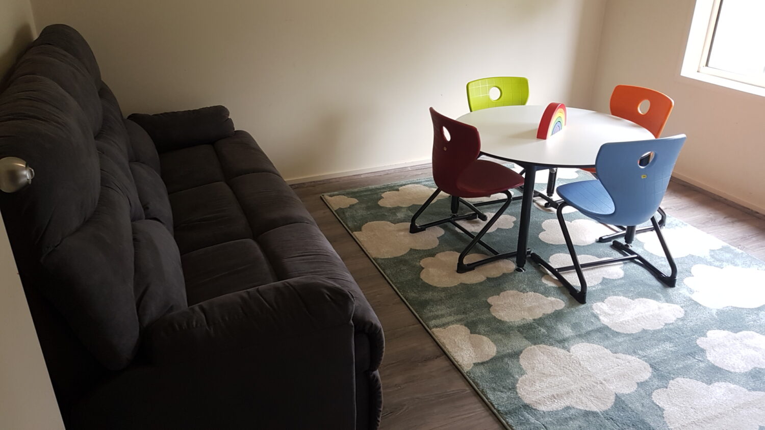 A cozy therapy space for rent features a black sofa, a round white table surrounded by red, green, blue, and orange chairs, and a cloud-patterned rug. Perfect for creating a calm and inviting atmosphere.