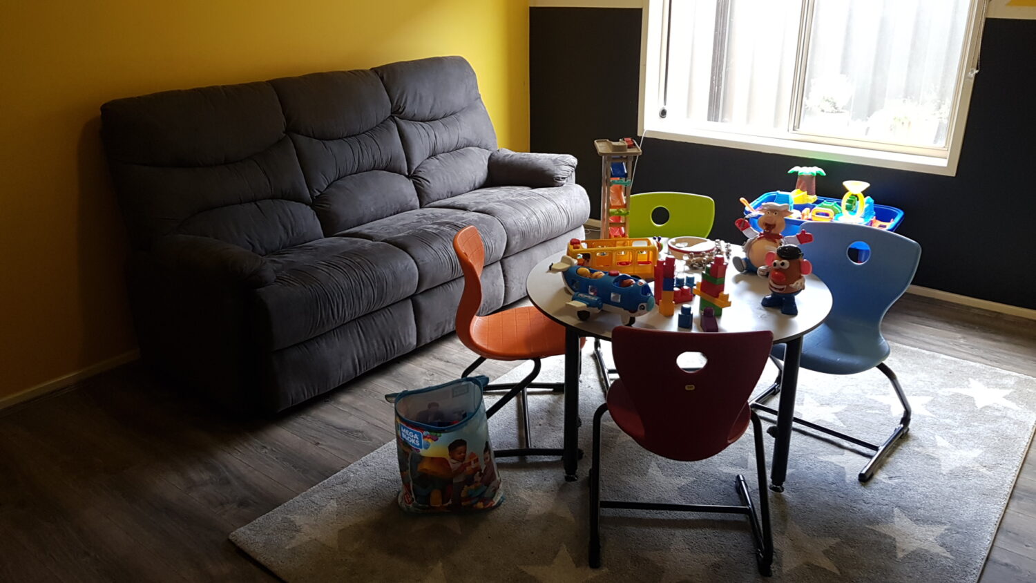 A children's playroom with a grey sofa, a round table with four multicolored chairs, and various toys scattered on the table and floor. A window illuminates the room, making it an ideal setting for therapy room rental due to its cozy atmosphere.