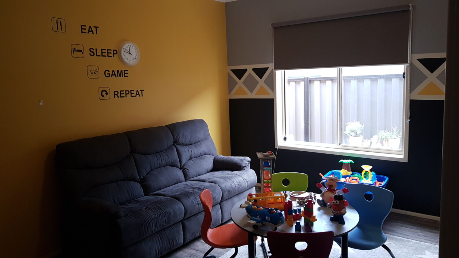 A cozy rental therapy space with a couch, toys on a small table, and colorful chairs. The wall has a clock and the phrase "eat sleep game repeat. " a window with a closed blind lets in some light.