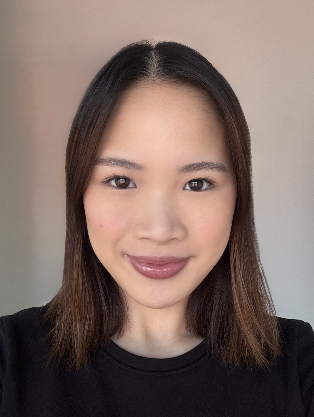 A person with straight, shoulder-length brown hair and natural makeup smiles slightly at the camera against a plain background.