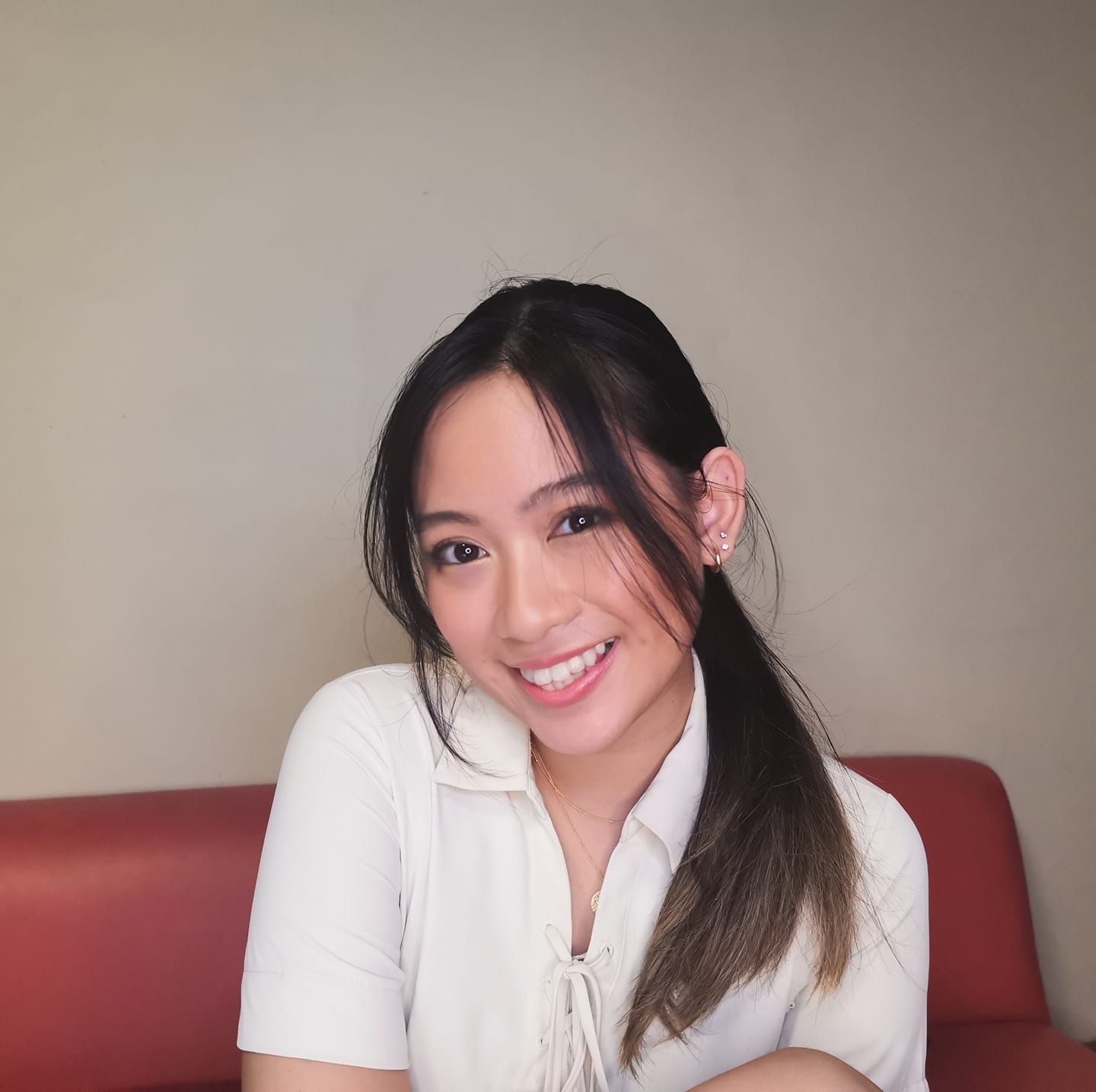 A young woman with long dark hair, smiling at the camera, wearing a white blouse, sitting on a red sofa with a beige wall in the background. She is Indiana Ramos.