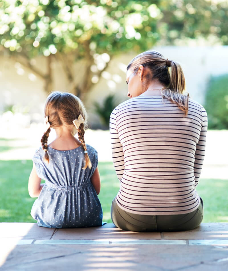 A woman and a young girl with pigtails sit on a porch step, facing away, looking at a garden with trees and greenery.