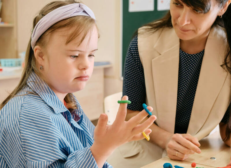 A young girl with Down syndrome looks at colored rings on her fingers, with an adult woman seated beside her at a table, observing attentively.