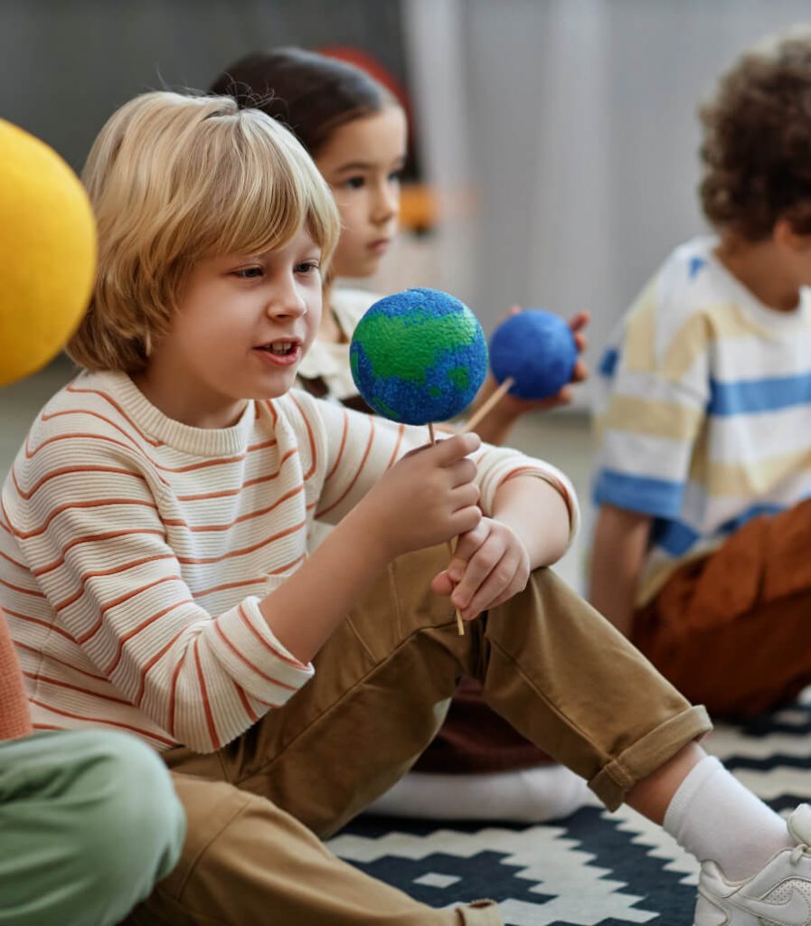 A child with blonde hair holds a model of the Earth while sitting on the floor. Other children are seated nearby, some holding similar models.