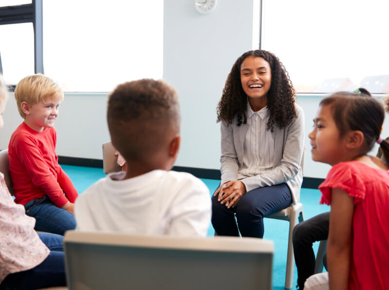 A woman sits on a chair, smiling and engaging in conversation with a group of children seated in a circle in a brightly lit room.