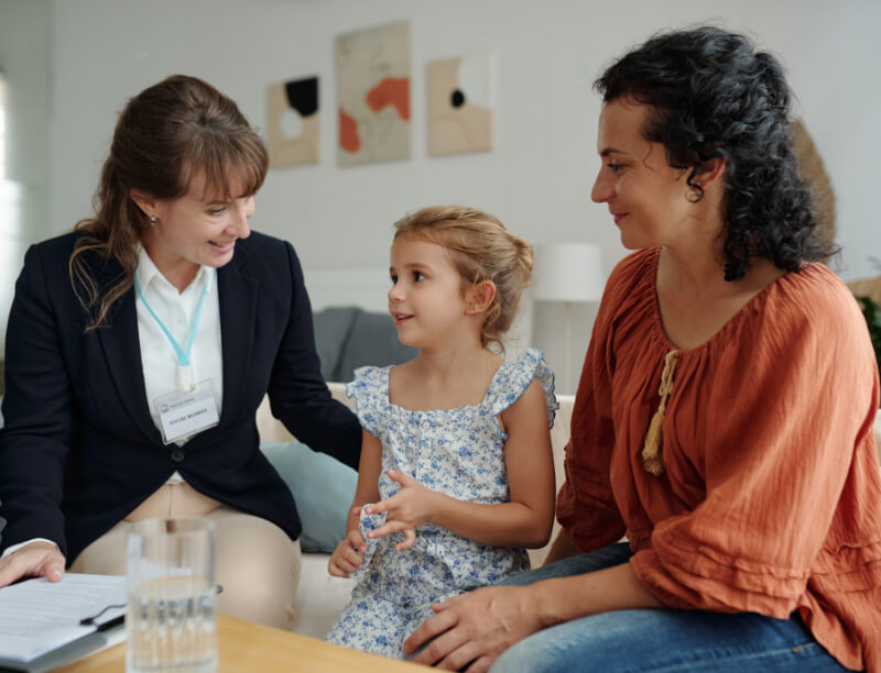 A woman in a blazer converses with a child and another woman, seated on a sofa. The child wears a floral dress. Papers and a glass are on the table in front of them.