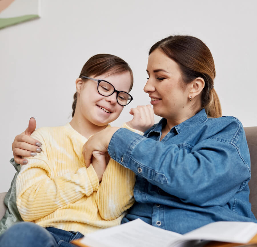 A person with glasses and a yellow sweater is happily sitting with another person in a denim shirt. They are both sitting on a couch with an open book in front.