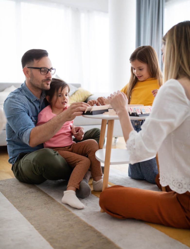 A family of four sits on the floor around a low table, playing a board game. The room is well-lit and cozy.