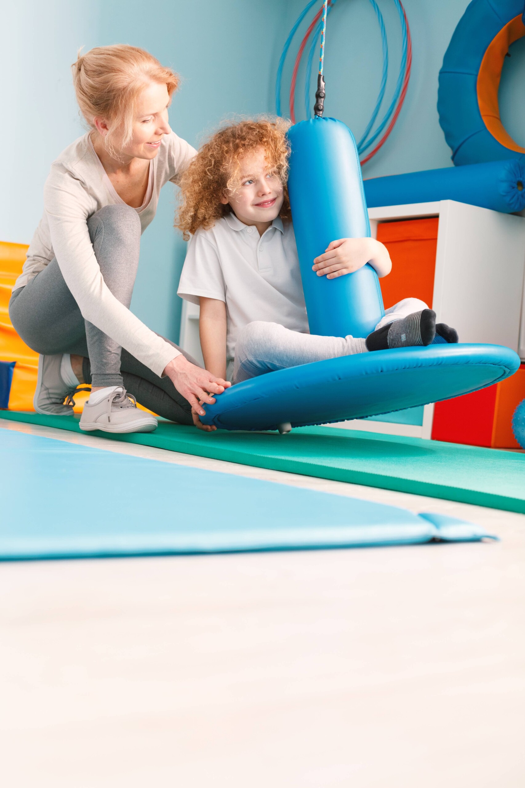 A child sits on a platform swing holding a blue cylindrical padding while an adult kneels beside, providing support. Colorful gym equipment can be seen in the background.