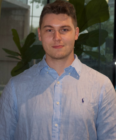 A man wearing a light blue button-down shirt stands indoors in front of a large plant and glass windows.