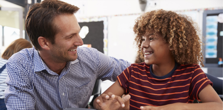 A man and a boy smiling at each other in a classroom.