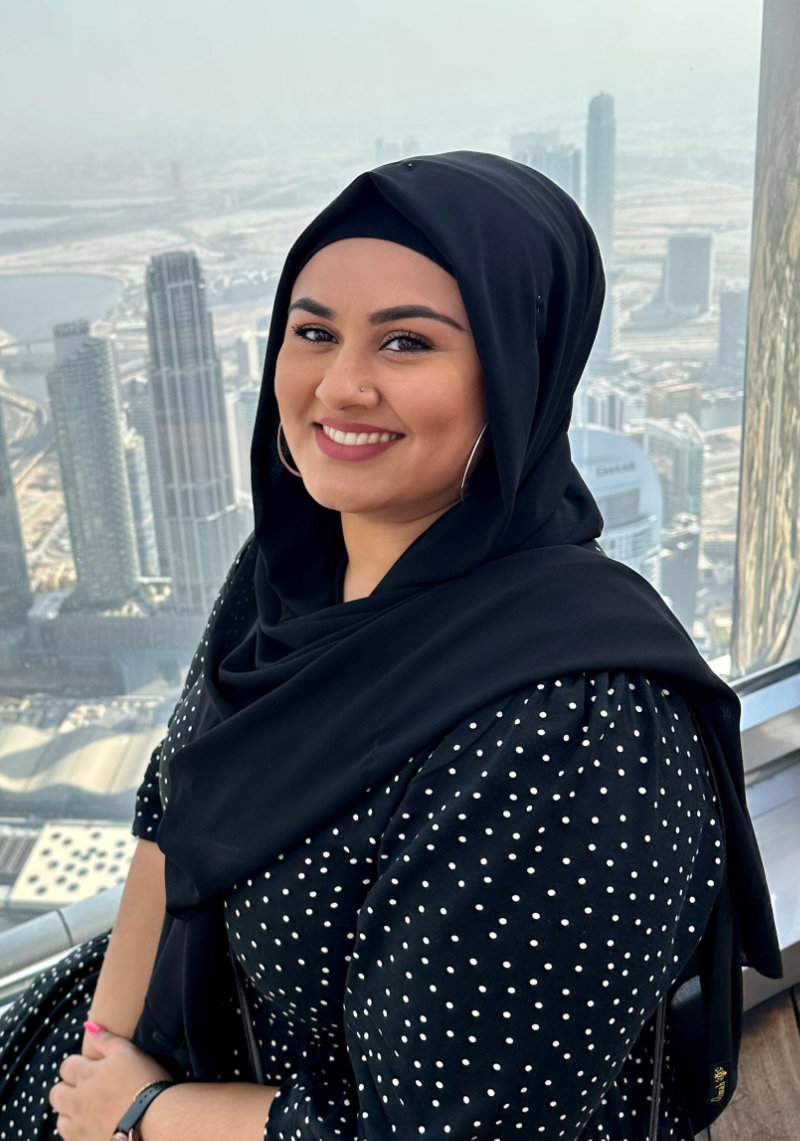 Zahrah, a hijab-wearing woman, smiling in front of a city view.