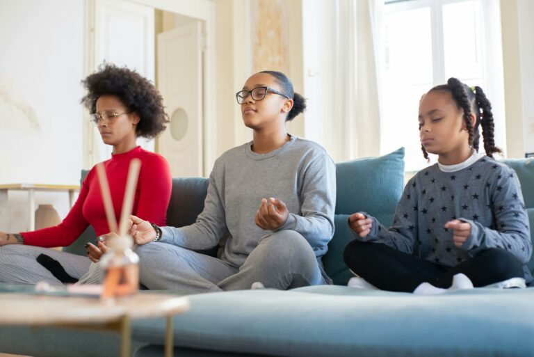 Three people are sitting on a couch in a calm meditation pose with their eyes closed and hands resting on their knees, practicing belly breathing to achieve calm and focus.