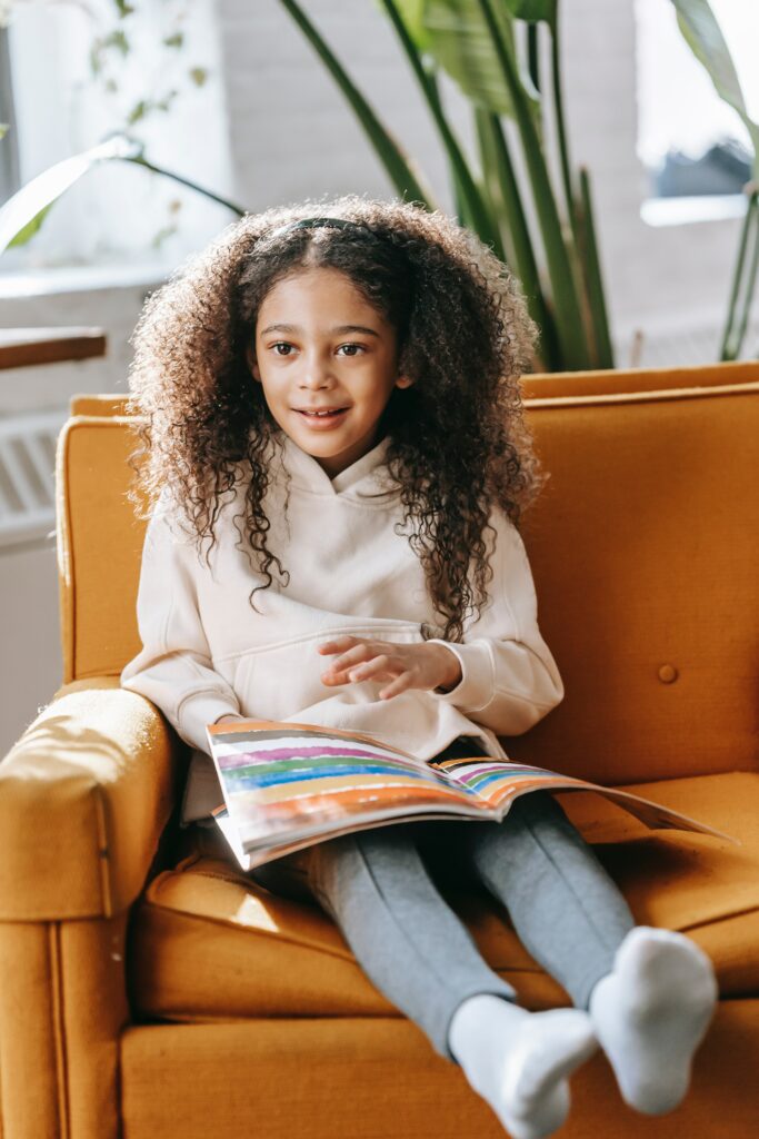 A little girl using the Picture Exchange Communication System (PECS) program sitting on an orange chair reading a book.