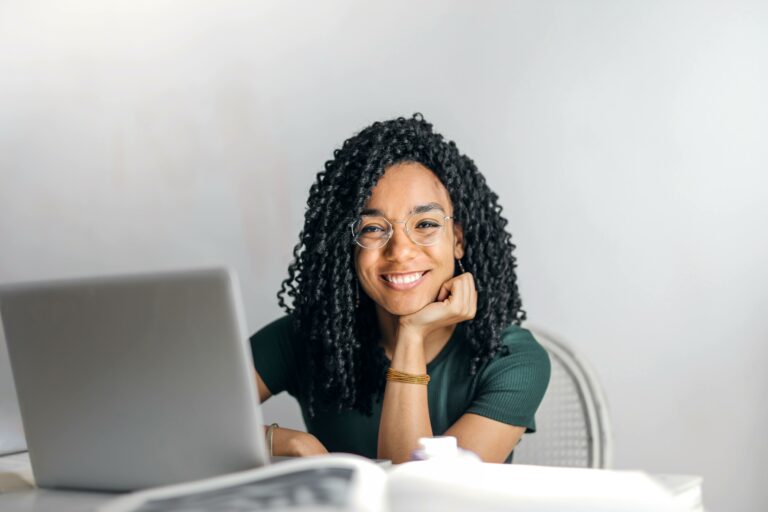 A person with curly hair and glasses smiles while sitting at a desk with an open laptop, accessing Quality NDIS services.