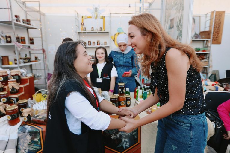 Two women smile and hold hands at an indoor market stall, discussing personal finance amid shelves of jars and other products. Other people stand nearby, adding to the lively atmosphere.