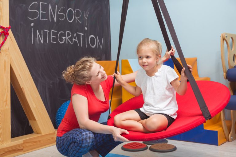 An adult supports a child sitting on a red swing in a therapy room, highlighting sensory processing. "Sensory Integration" is written on a chalkboard in the background.