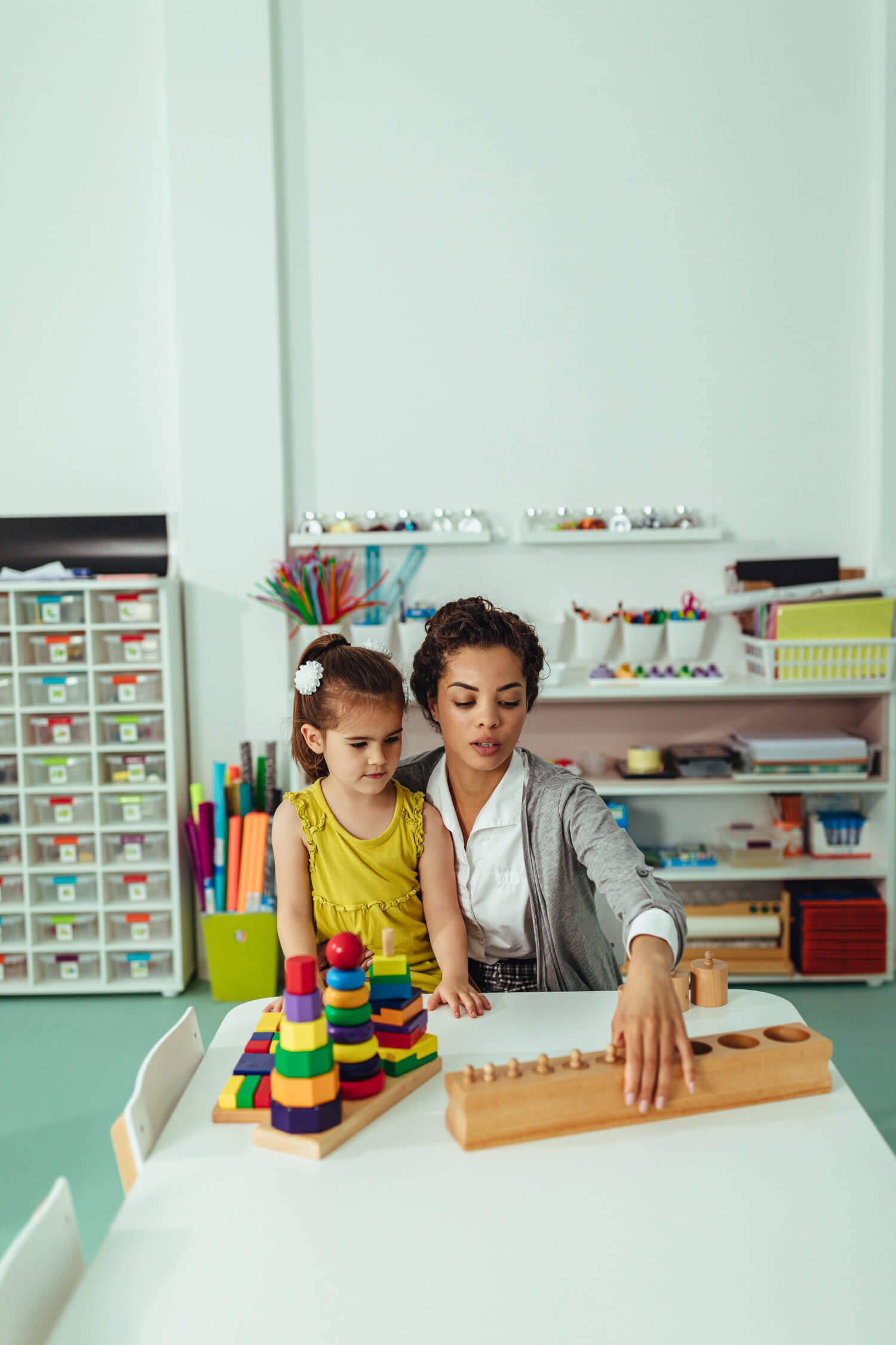 A teacher assists a young student with educational toys in a classroom. The room is equipped with various learning materials and storage shelves.