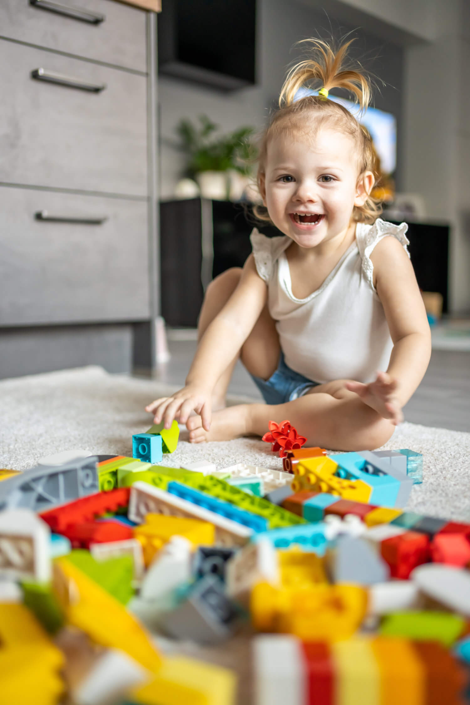 A smiling toddler with a ponytail sits on a carpet indoors, playing with colorful building blocks spread out in front of them.
