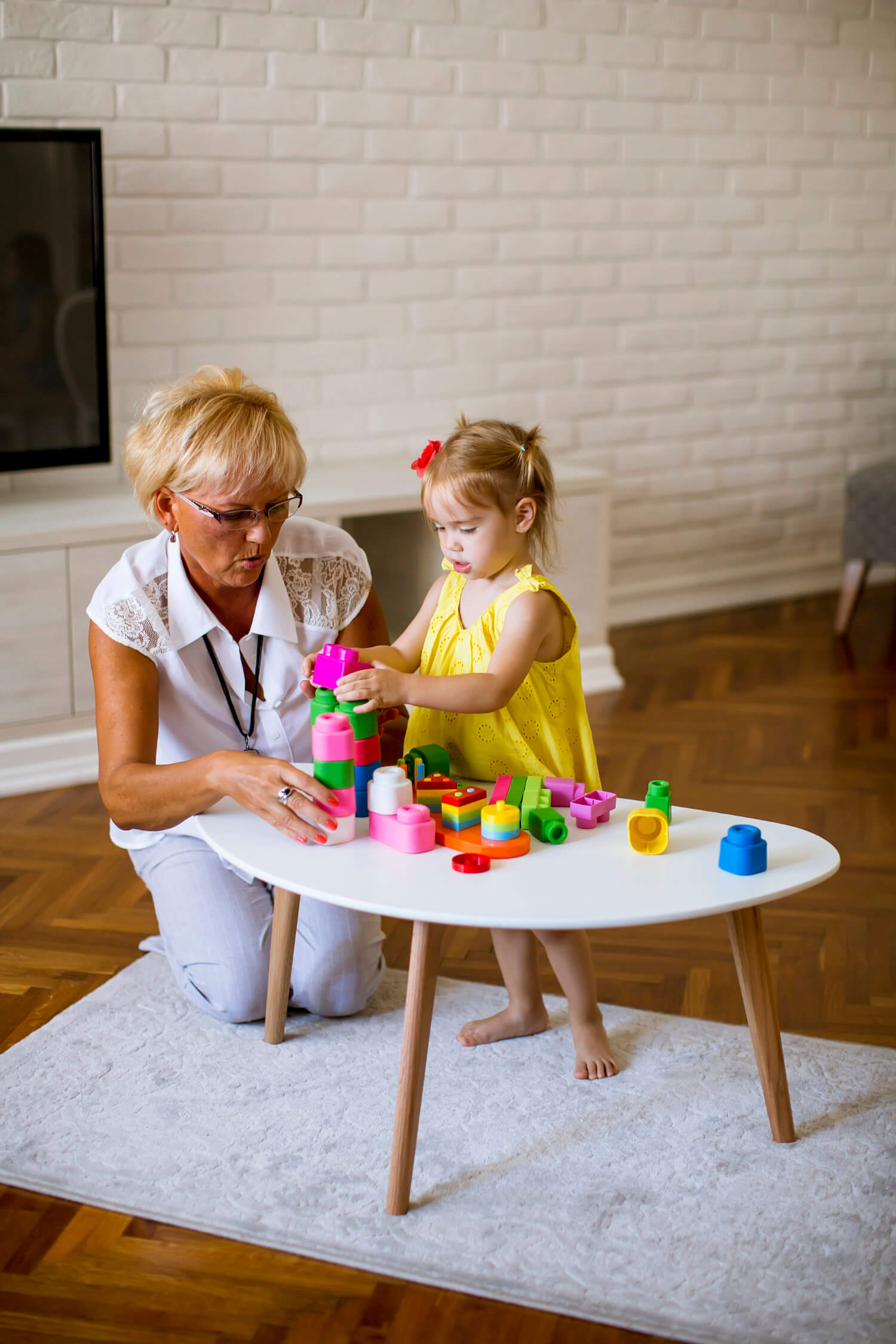 An older woman and a young girl are playing with colorful stacking toys on a white table in a living room with a brick wall and wooden floor.