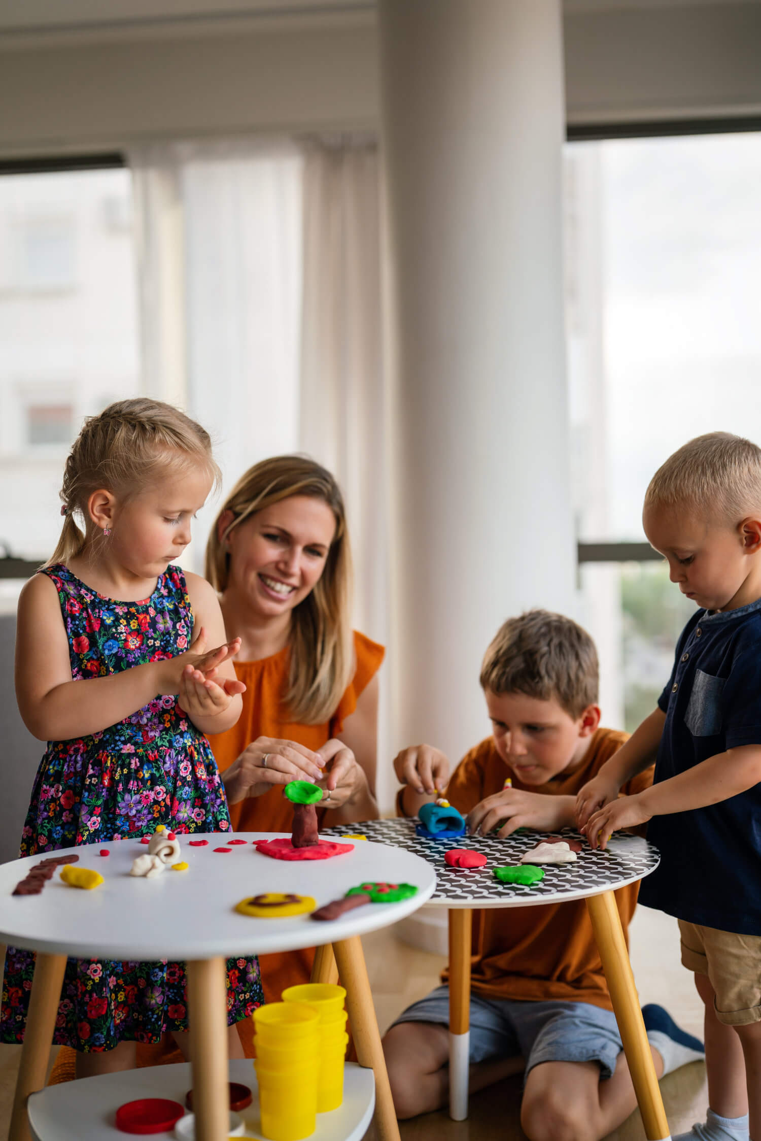 A woman and three children are sitting and standing around a small table, playing with colorful modeling clay in a bright room.